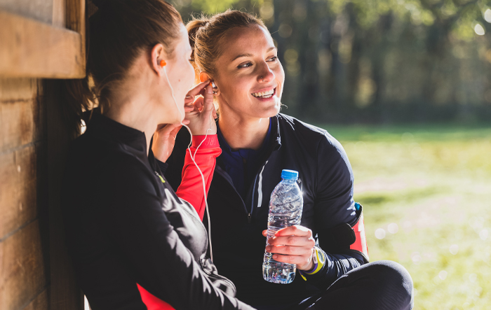 szczesliwa-sportsmenka-sluchanie-muzyki-i-trzyma-butelke Dwie uśmiechnięte kobiety po treningu słuchają muzyki i trzymają butelkę wody, odpoczywając na świeżym powietrzu
