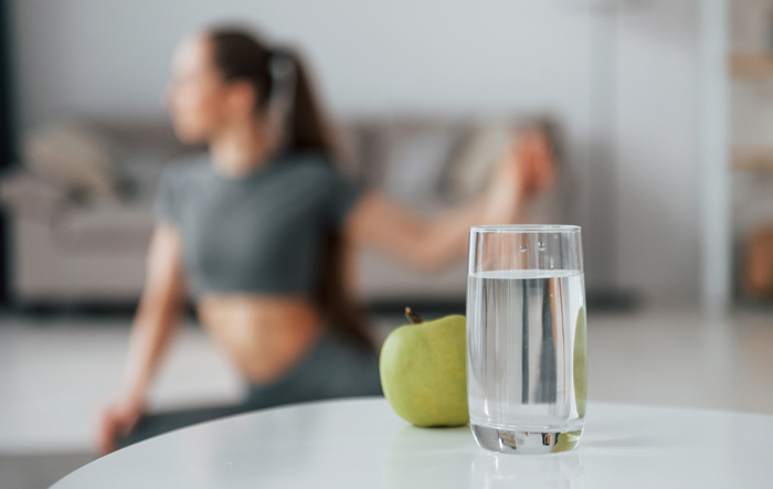 Szklanka wody i jabłko na stole, w tle osoba ćwicząca w domu – nawadnianie i dieta bez cukru