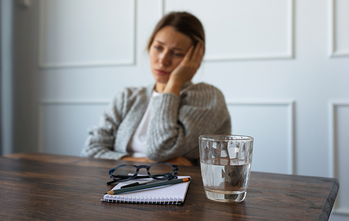 Kobieta siedząca przy stole z głową opartą na dłoni, obok szklanka wody &ndash; objawy lekkiego odwodnienia i zmęczenia.