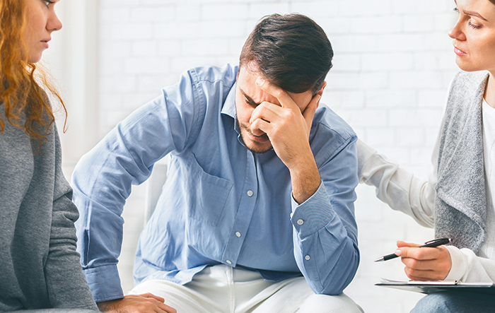 Mężczyzna siedzący z pochyloną głową w towarzystwie dw&oacute;ch os&oacute;b &ndash; stres i napięcie mogą wpływać na odczuwanie pragnienia.