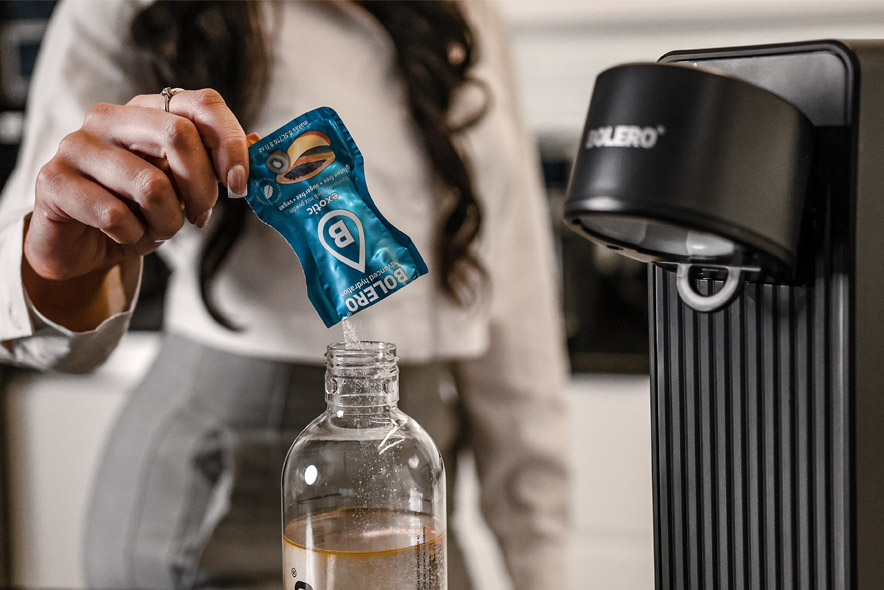 Kobieta wsypuje saszetkę Bolero do butelki z wodą przed przygotowaniem napoju w saturatorze Bolero Soda Maker.