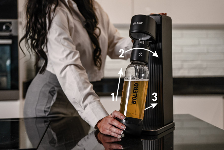 Instrukcja montażu: butelka z napojem jest unoszona i przekręcana, aby poprawnie zamocować ją w saturatorze Bolero Soda Maker.