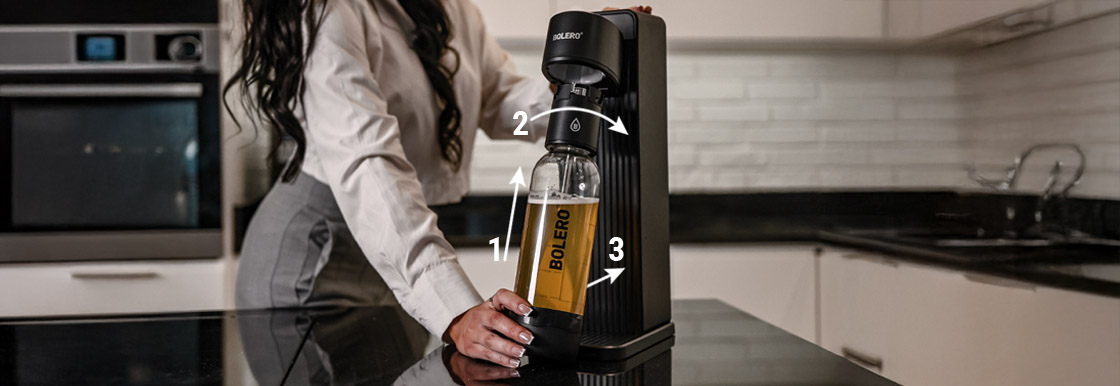 Instrukcja montażu: butelka z napojem jest unoszona i przekręcana, aby poprawnie zamocować ją w saturatorze Bolero Soda Maker.