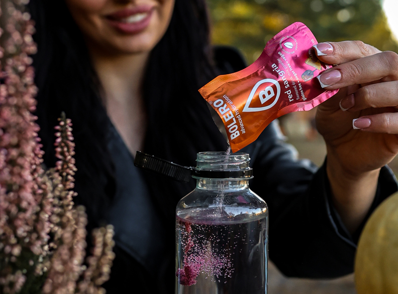 Bolero Red Orange – saszetka napoju o smaku czerwonej pomarańczy obok butelki z gotowym napojem, bez cukru, gluten free i vegan.