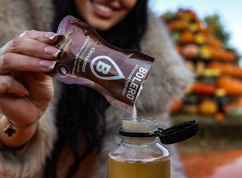 Przygotowanie napoju Bolero Cinnamon – wsypywanie saszetki z cynamonowym napojem bez cukru do butelki z wodą, jesienny klimat z dyniami w tle.