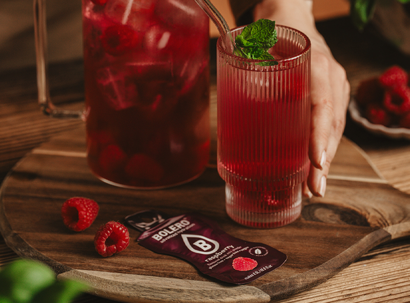Bolero Raspberry – napój o smaku malinowym bez cukru, w szklance z listkiem mięty i świeżymi malinami, obok saszetka produktu.